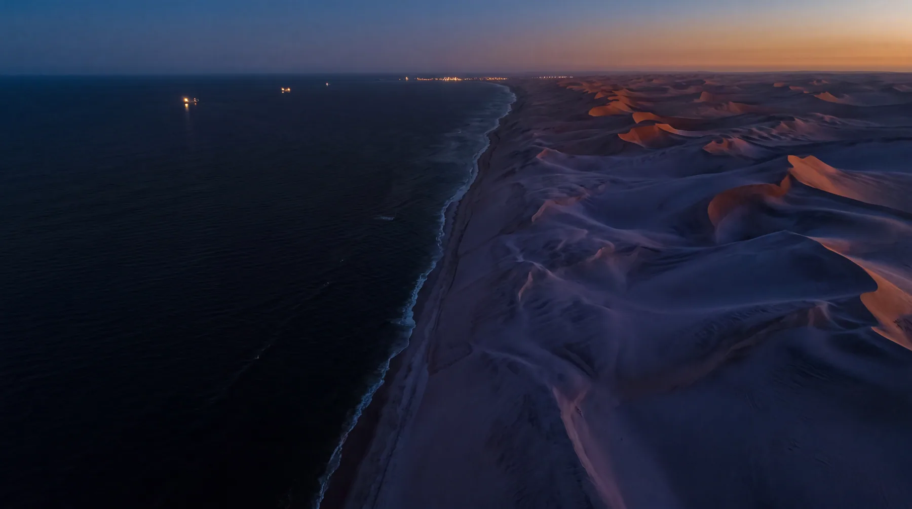 Aerial view of Namib dunes meeting the Atlantic coast at dusk, with ship lights offshore.